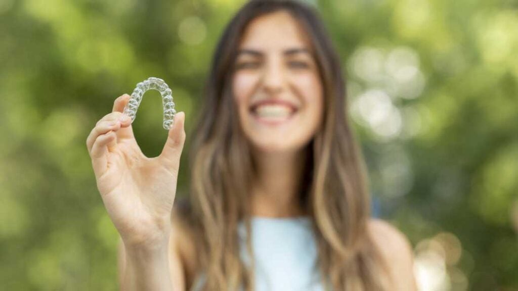 Smiling young woman holding a clear Invisalign aligner outdoors